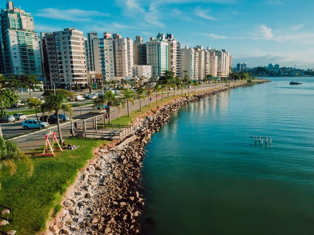 Vista aérea da orla urbana com parque, prédios residenciais à beira da água, área verde para lazer e avenida costeira com circulação de veículos.