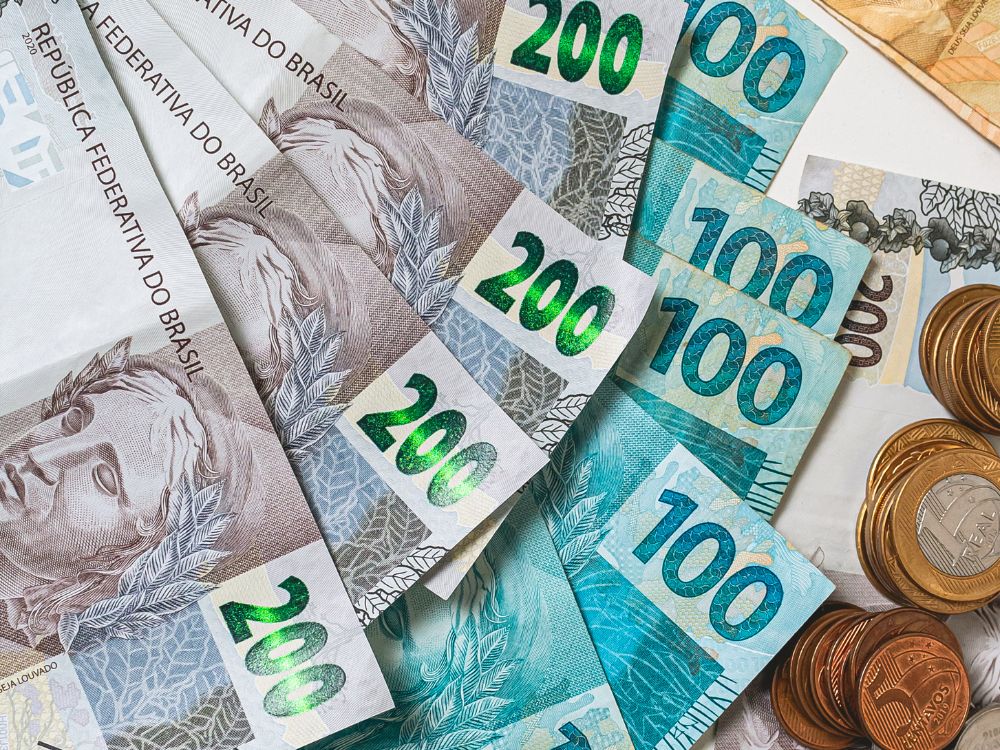 Cédulas de real de 200 e 100 reais espalhadas sobre superfície clara, acompanhadas de moedas brasileiras, representando organização e controle financeiro.