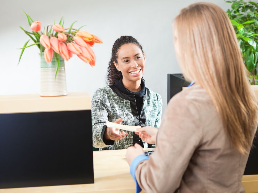 Atendente sorrindo entrega cartão ou documento a cliente em balcão de atendimento, representando contato e orientação sobre como falar com o jurídico do Bradesco.